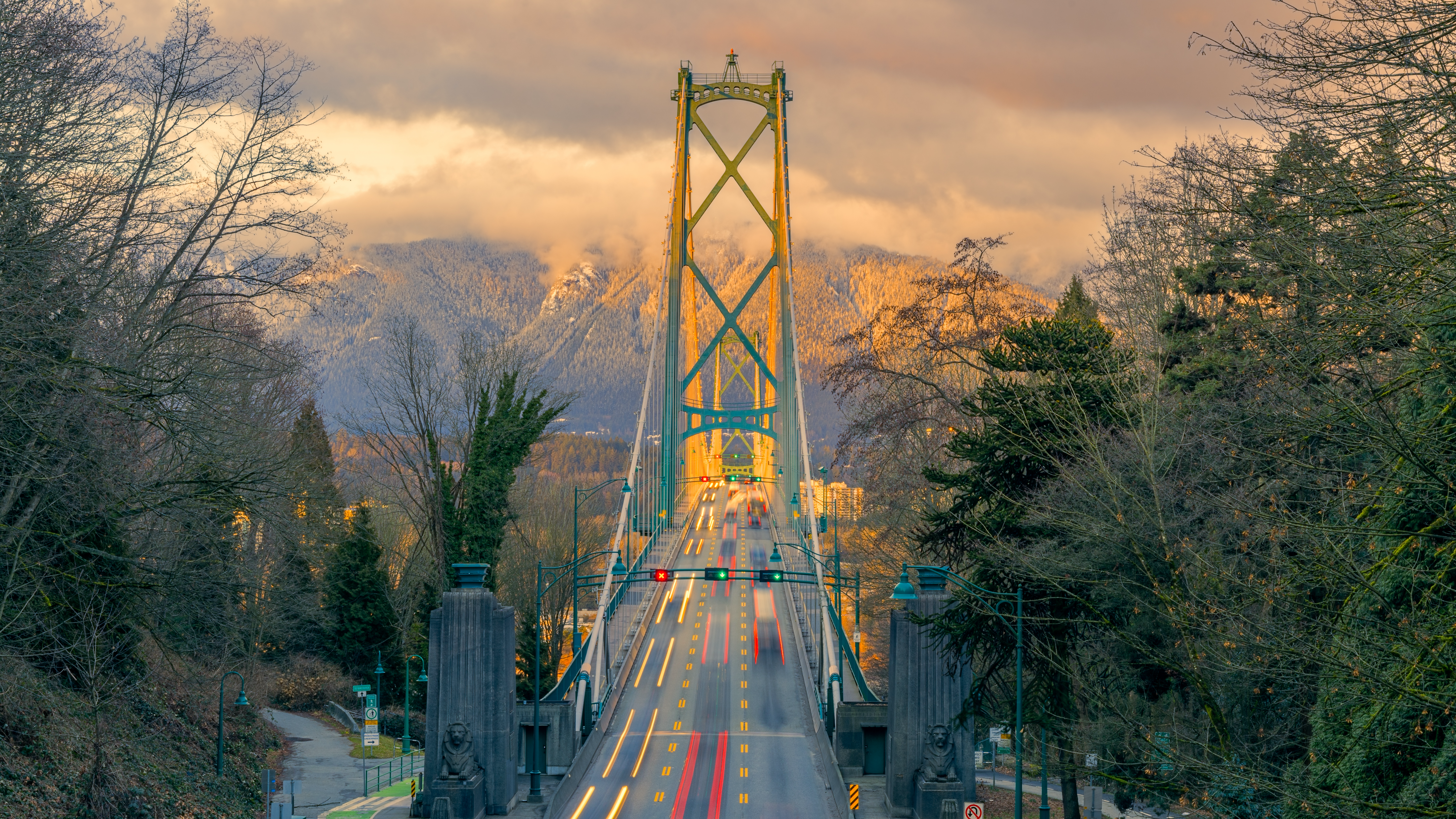 Scenic view of the Lions Gate Bridge in Vancouver, BC, spanning across Burrard Inlet, surrounded by lush greenery and distant mountain peaks under a clear blue sky.
